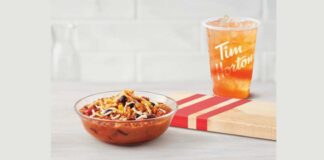 Tim Horton hearty loaded bowl of chili in front of a wooden cutting board with a cup of ice tea behind cutting board