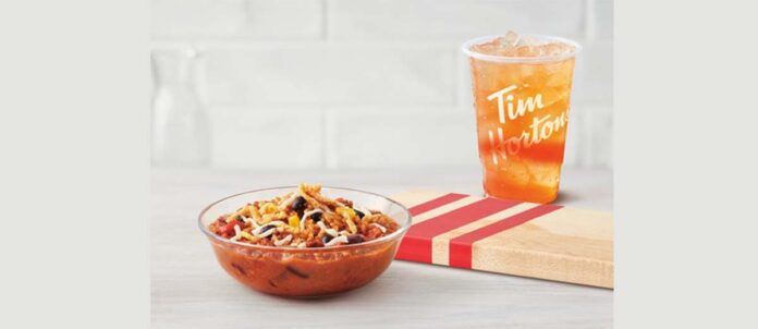 Tim Horton hearty loaded bowl of chili in front of a wooden cutting board with a cup of ice tea behind cutting board