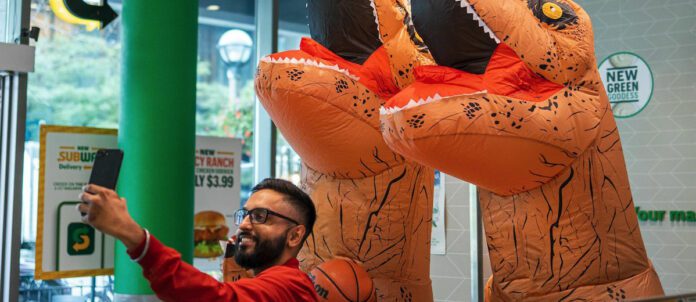 Man taking a photograph at a Subway with Toronto Raptors inflatable mascots behind him