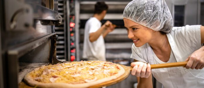 Female chef in white uniform and hairnet putting pizza in the oven