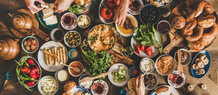 Flat-lay of peoples hands taking pastries, vegetables, greens, olives, cheeses, fried eggs, spices, jams, honey, tea in copper pot and tulip glasses, wide composition