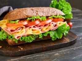 submarine sandwich with ham, cheese, lettuce, tomatoes and microgreens on old wooden dark background