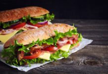 Two fresh submarine sandwiches with ham, cheese, bacon, tomatoes, lettuce, cucumbers and onions on dark wooden background