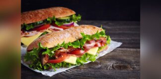 Two fresh submarine sandwiches with ham, cheese, bacon, tomatoes, lettuce, cucumbers and onions on dark wooden background