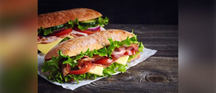 Two fresh submarine sandwiches with ham, cheese, bacon, tomatoes, lettuce, cucumbers and onions on dark wooden background